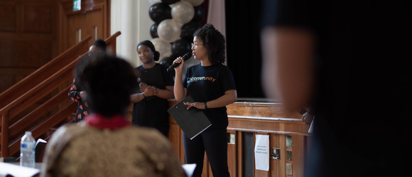 Woman leading community event with microphone against backdrop of Black Thrive branding and black and white balloons