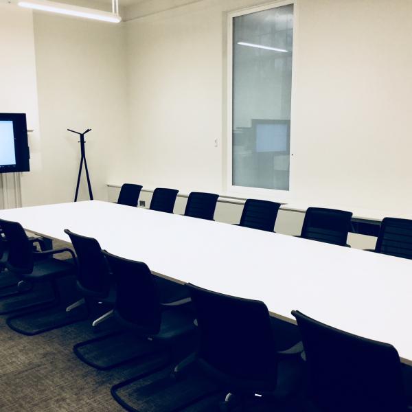 Venue image of white desks in boardroom style with black chairs around them with television screen at the back of the room
