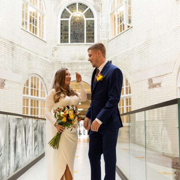 Couple standing on glass walkway in brick building with large arched windows, with bride holding bouquet flowers 