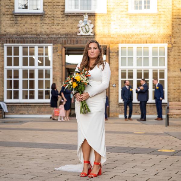 Bride stands in Lambeth Town Hall Courtyard wearing white dress, holding yellow flowers 