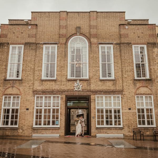 External facade of Lambeth Town Hall and outside courtyard area, couple walking through main doors in the rain with umbrella